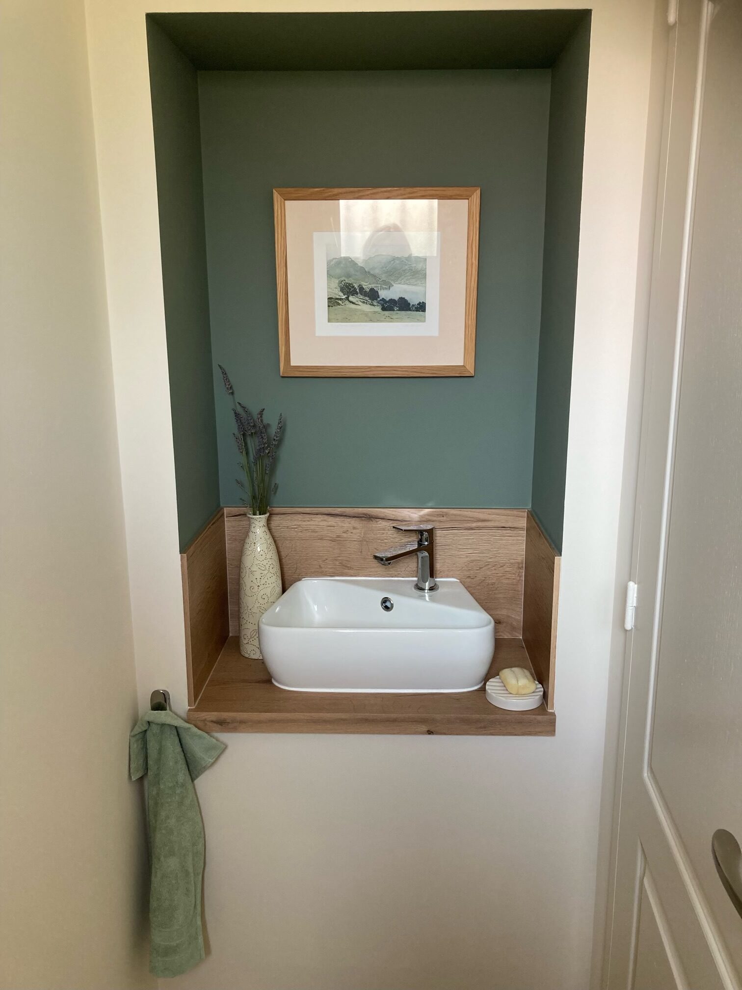 Vasque en céramique posée sur un meuble en bois clair, dans une niche murale peinte en vert.