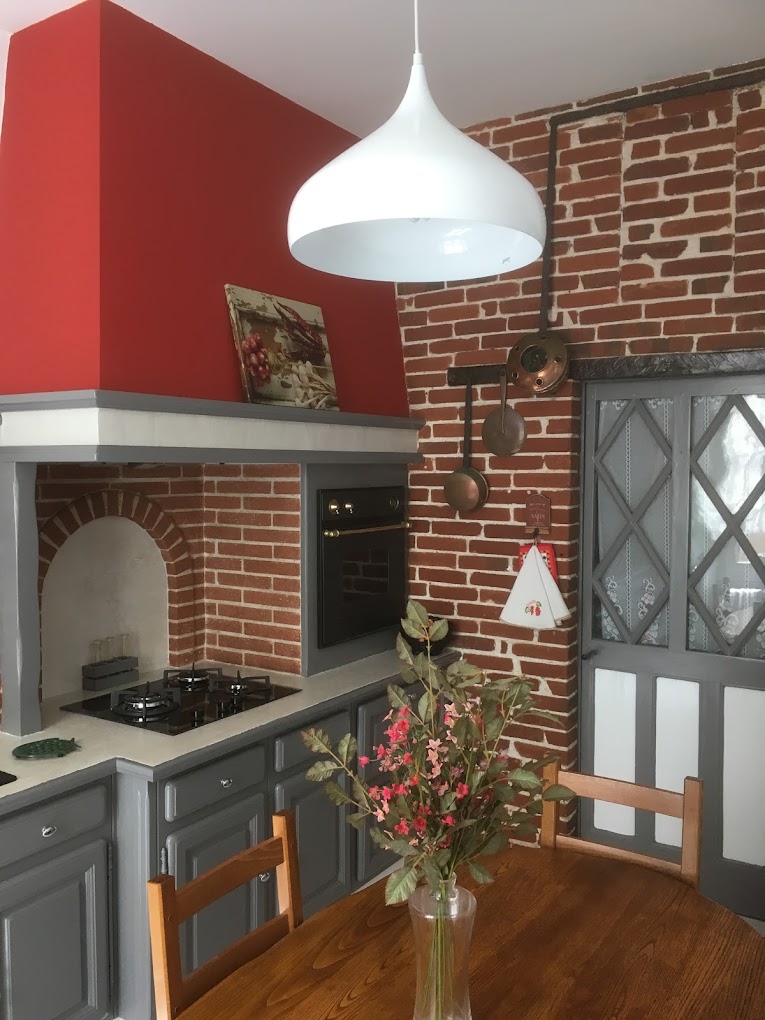 Cuisine ouverte sur la salle à manger, avec une cheminée en briques rouges qui apporte une touche chaleureuse et authentique. Les armoires grises contrastent élégamment avec les murs rouges et la cheminée. Un plan de travail en pierre naturelle ajoute une note de rusticité