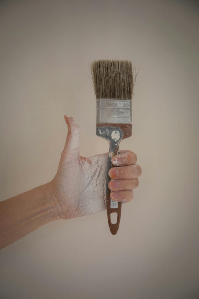 A paint-covered hand holding a paintbrush in front of a white wall, signifying creativity.